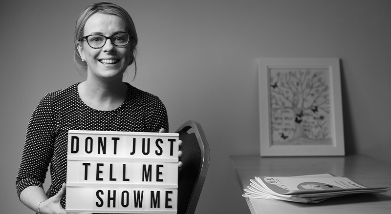 Black and white image of Catriona Watters Crehan holding a sign 'Don't just tell me , show me'